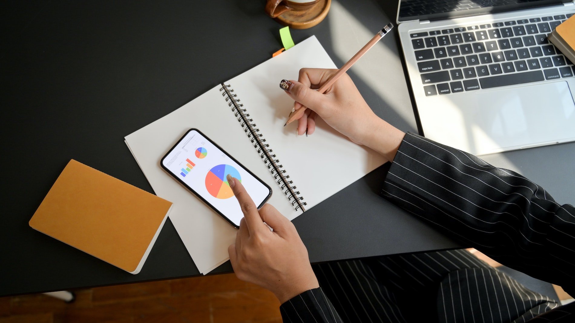 Konzepte und digitale Marketing Strategien - Frau am Arbeitsplatz mit einem Notizbuch und Stift in der Hand. Daneben ein ein Handy mit einem Kreisdiagramm. Rechts daneben ein offener Laptop.